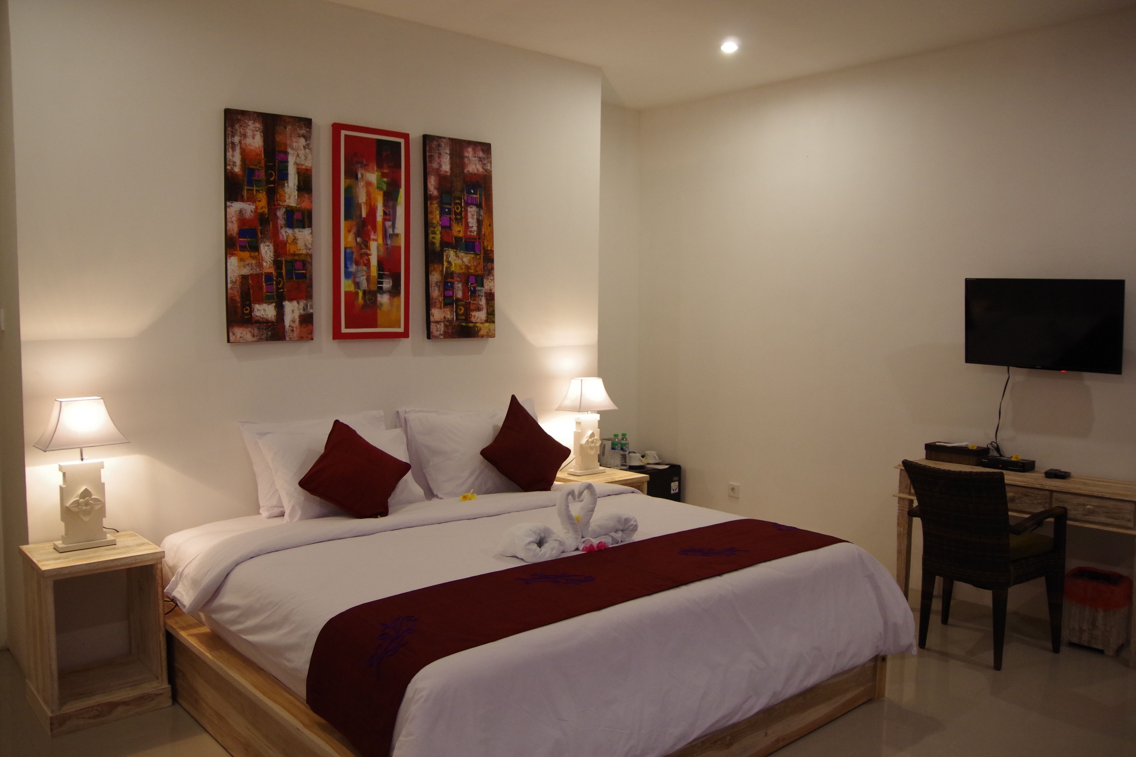 A hotel room with a large bed, two bedside tables with lamps, a desk with a chair, a flat-screen TV, and three abstract paintings on the wall.