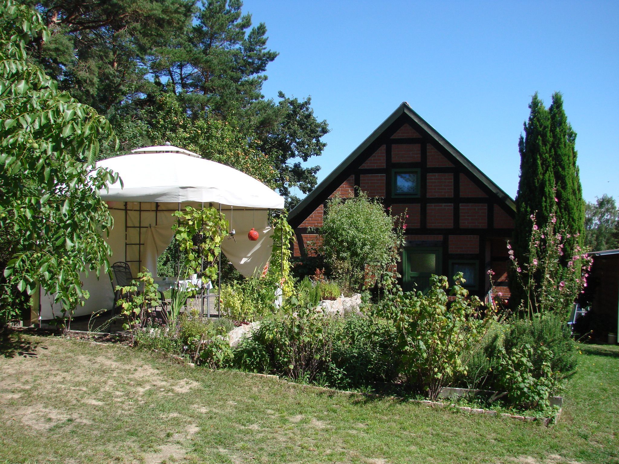 Ein traditionelles Haus mit geneigtem Dach, umgeben von einem gepflegten Garten mit verschiedenen Pflanzen und einer weißen Markise.