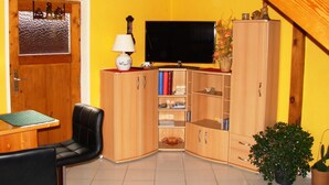 Una habitación con puerta de madera, televisor sobre un mueble, un aparador con libros, una mesa con una silla y una planta en maceta.