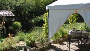 Un patio jardín con una carpa blanca, una mesa con sillas y un edificio pequeño con techo de tejas.