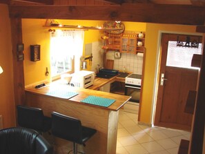A kitchen with wooden cabinets, a microwave, and a stove. There are two black bar stools and a window with curtains. - Vacation cottage Bildt, Germany (Loddin)
