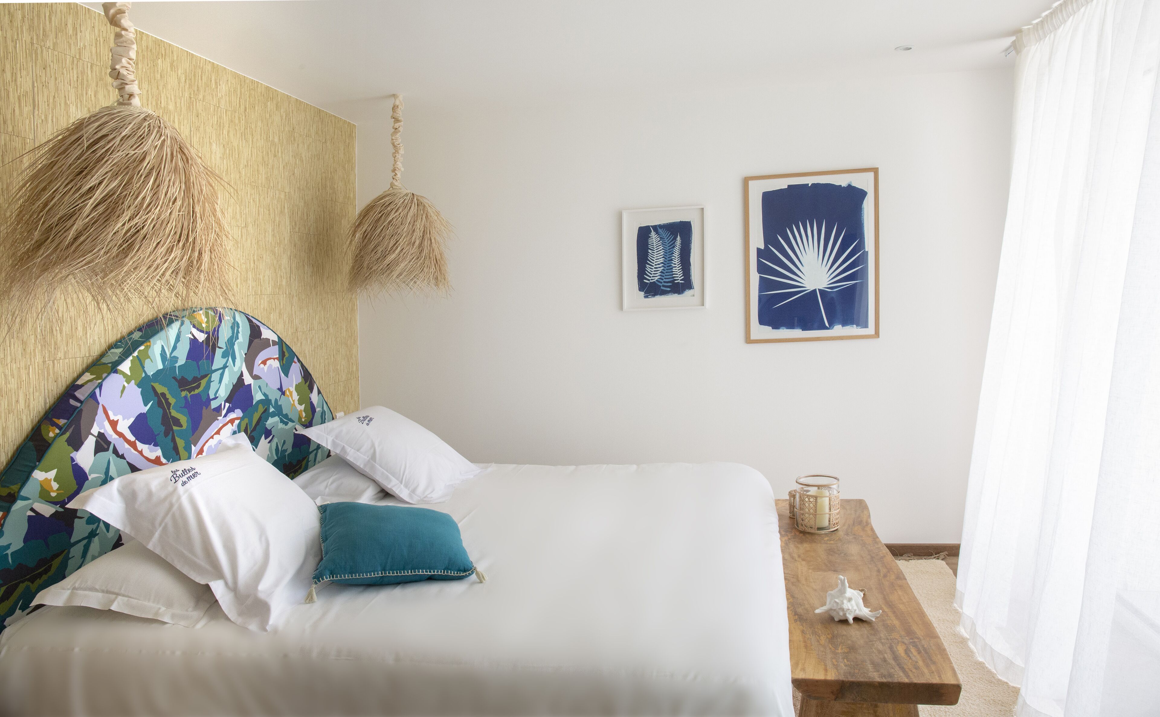 Una cama bien hecha con almohadas blancas y azul verdosas, un cabecero tejido y dos cuadros enmarcados en la pared.