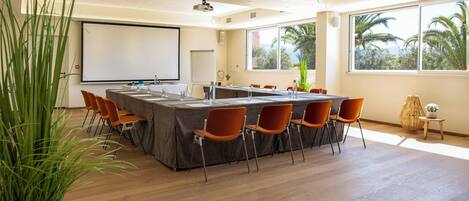 Un salón de conferencias con una larga mesa, sillas anaranjadas, una pantalla de proyector y ventanas amplias con vista a palmeras.