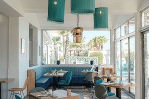 An indoor seating area with tables and chairs, a bench, and a view through large windows. - Les Bulles de Mer (Saint-Cyprien)