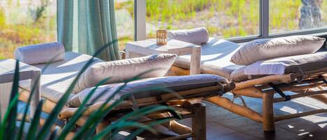 A sunlit room with wooden lounge chairs, white cushions, and a small table with a candle.