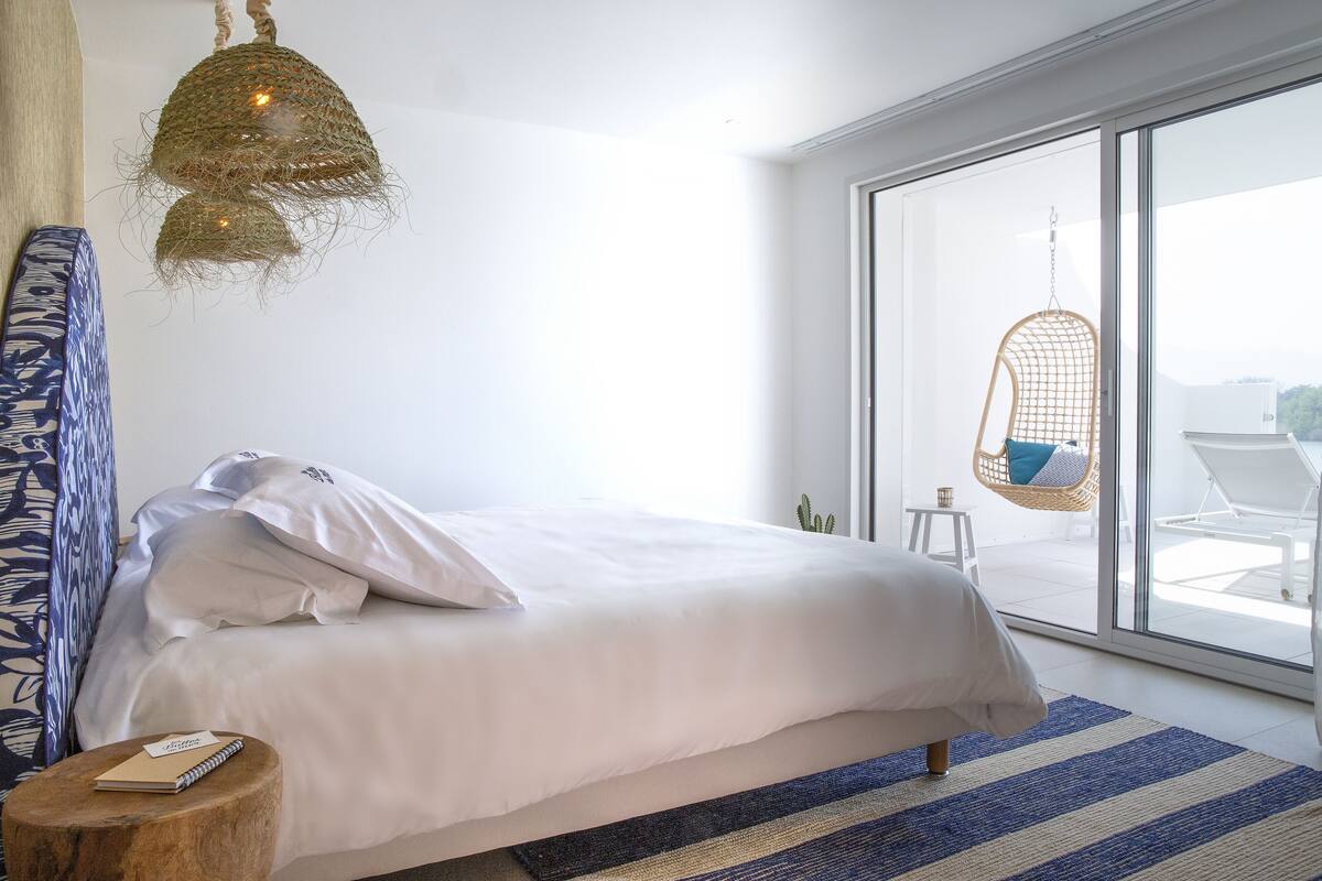 Une chambre à coucher moderne, dotée d’un grand lit, d’un luminaire suspendu et offrant une vue sur l’extérieur grâce à une porte vitrée.