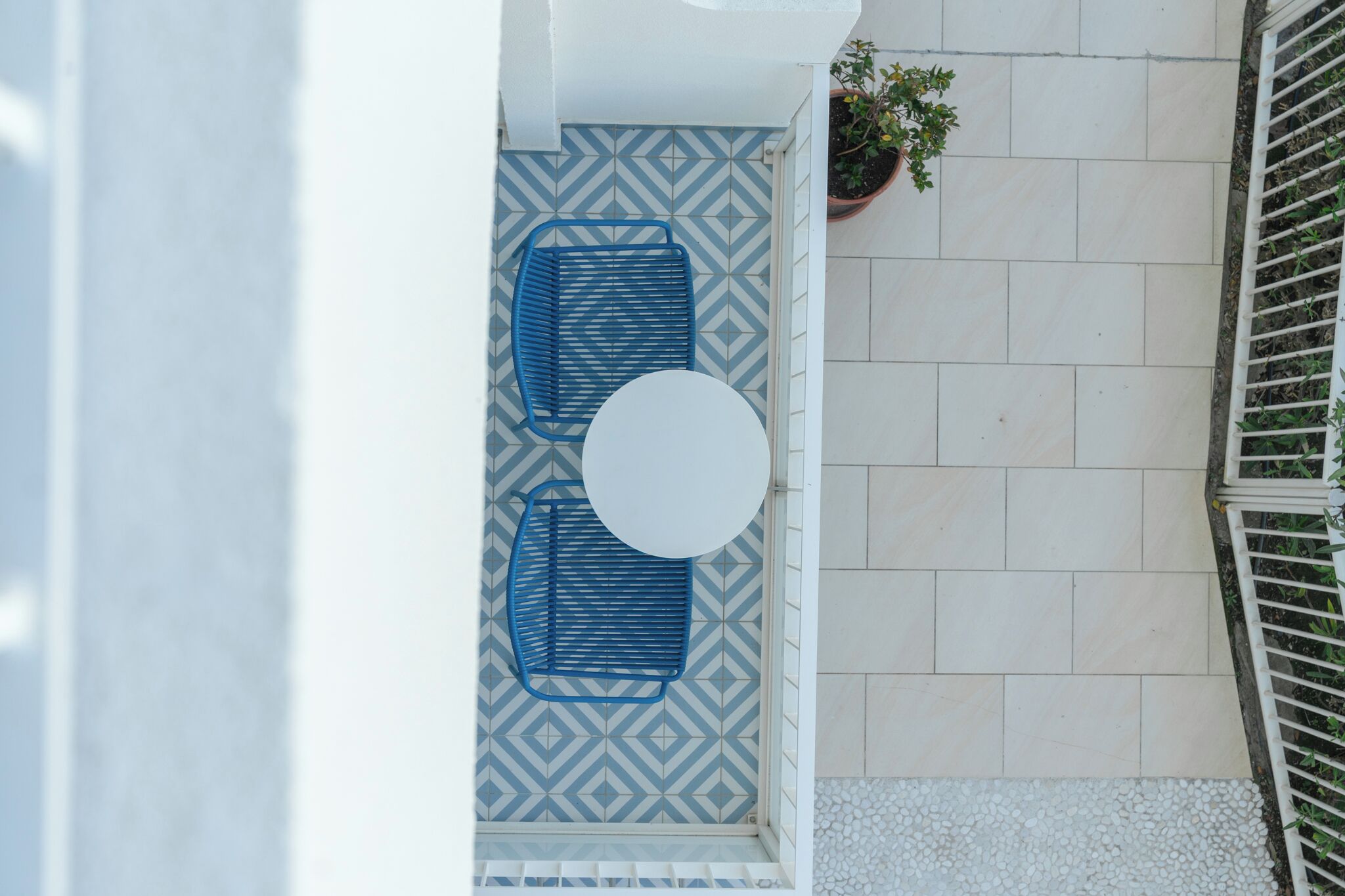 A patio with a white round table and two blue chairs, a potted plant, and a patterned floor.