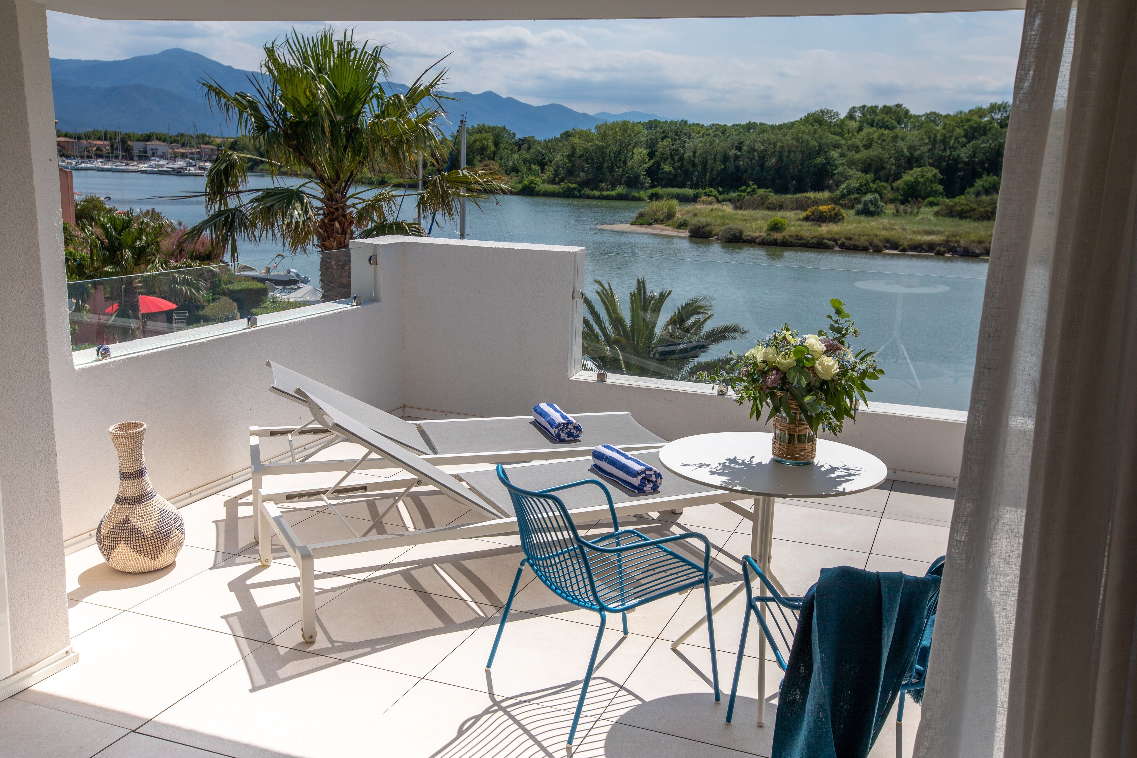 A balcony with a view of a river and mountains, equipped with two lounge chairs, a small table, and a vase.