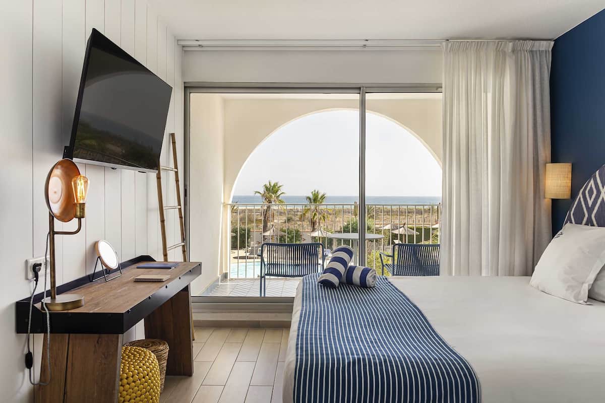 Une chambre d’hôtel comprenant un lit, un bureau avec un téléviseur à écran plat, une vue sur l’océan et un balcon aménagé avec du mobilier d’extérieur.
