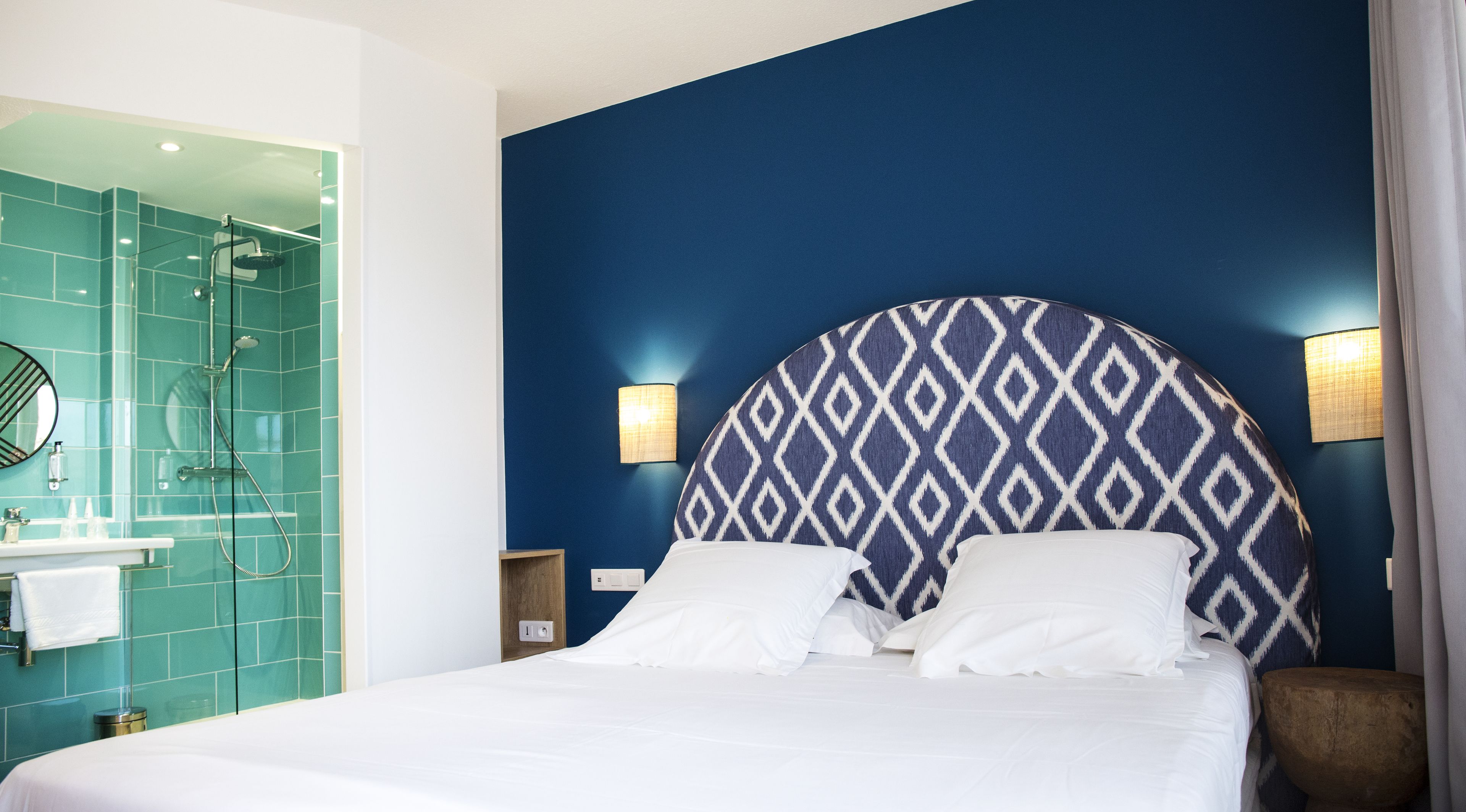A hotel room with a bed, a bathroom with a shower, and a wall with a patterned headboard.