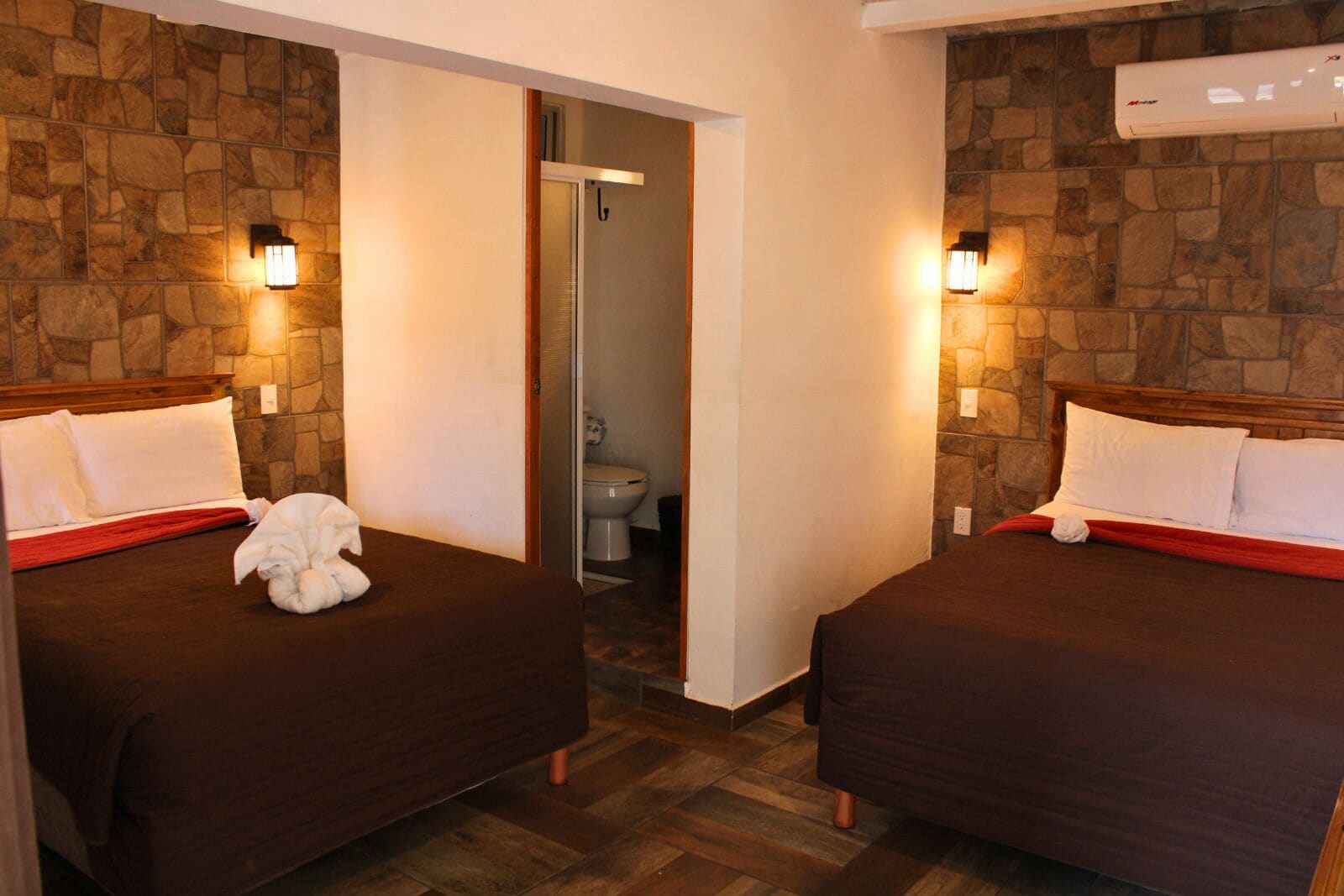 Habitación de hotel con dos camas, una pared de piedra, piso de madera y un baño visible a través de una puerta abierta.