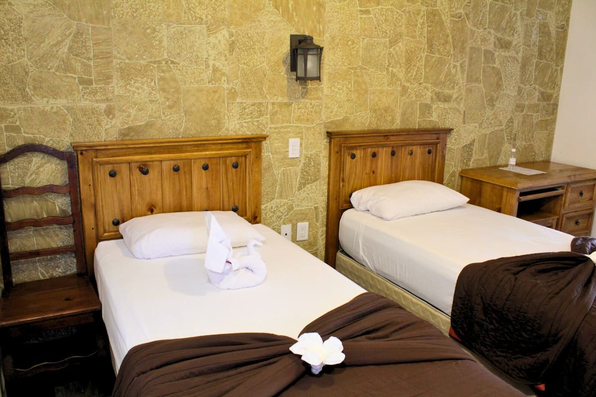 Een hotelkamer met twee bedden, houten hoofdeinden, stenen muren, een nachtkastje en een lamp.