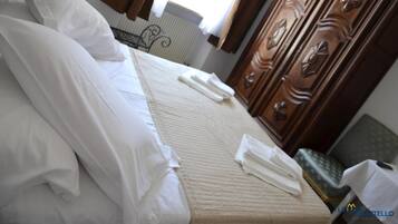A neatly made bed with white linens and towels, a wooden wardrobe with decorative handles, and a radiator in the background.