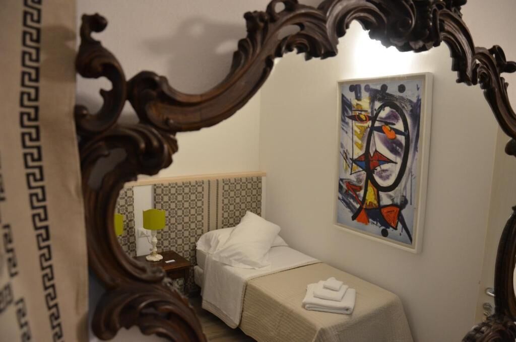 A hotel room with a bed, a small table with a lamp, a painting on the wall, and an ornate mirror frame.