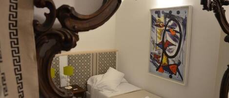 A hotel room with a bed, a small table with a lamp, a painting on the wall, and an ornate mirror frame.