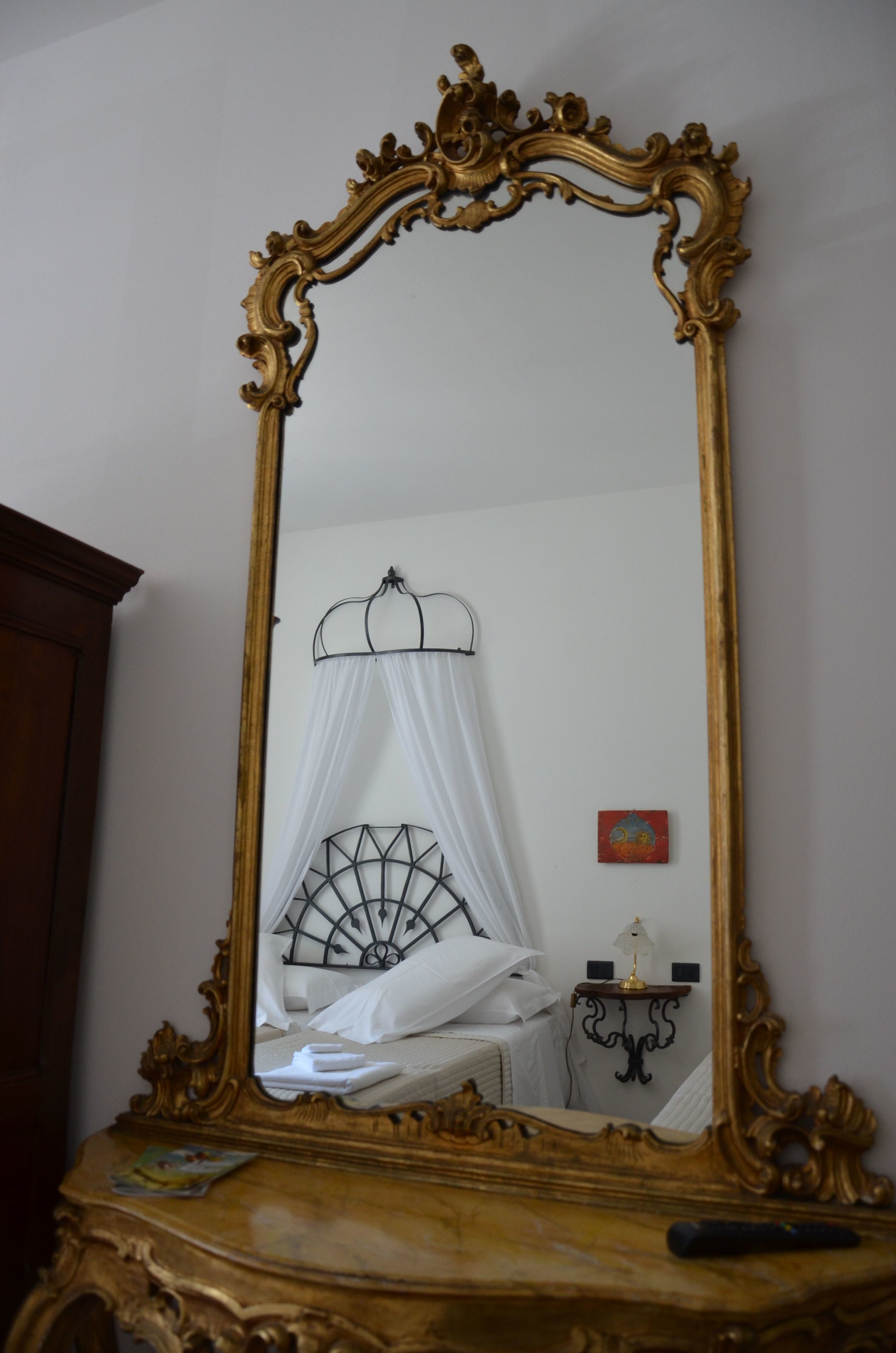 A bedroom with a large ornate mirror on a wooden dresser, a bed with a white canopy, a small bedside table with a lamp, and a wall-mounted artwork.