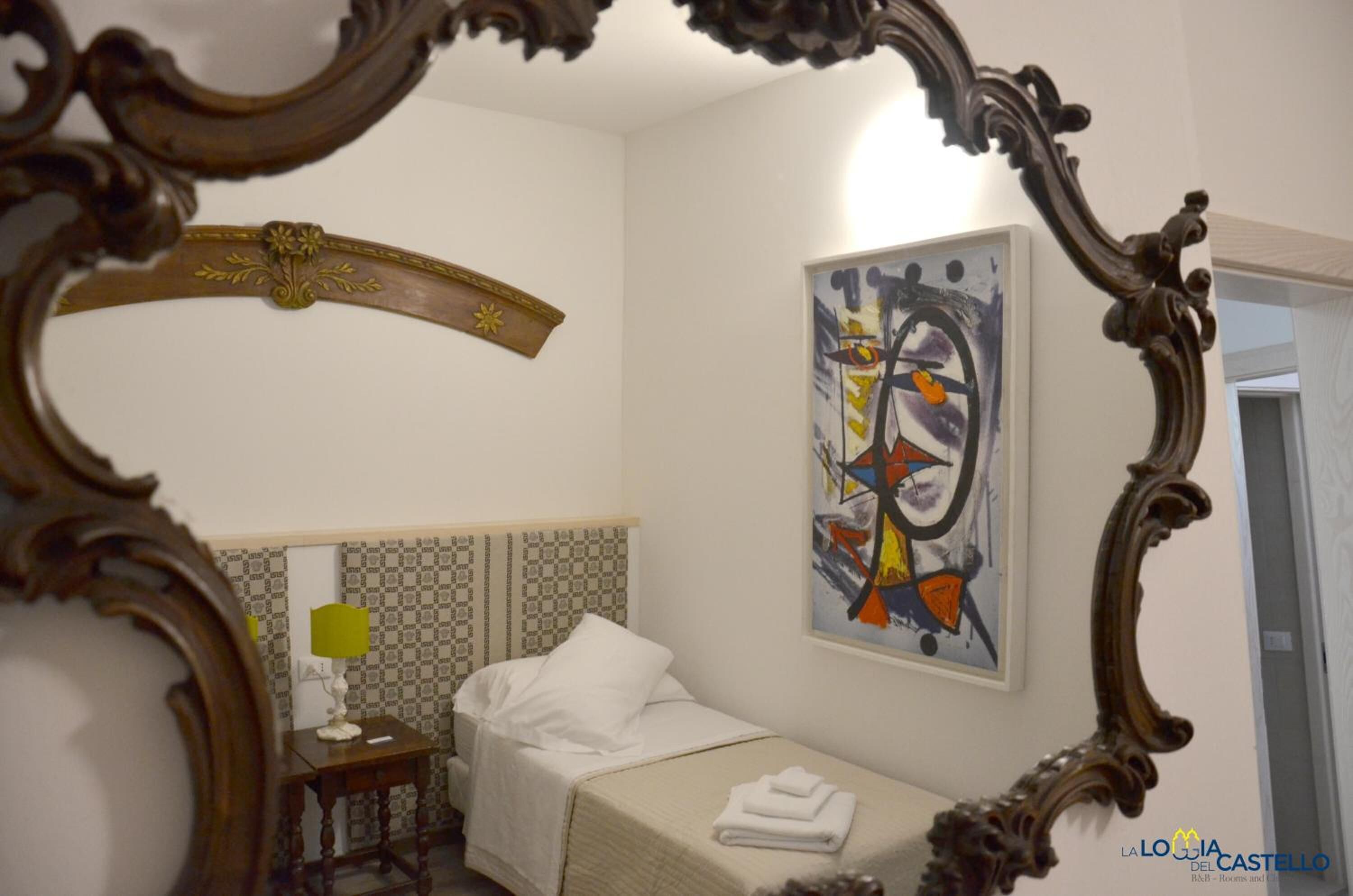 A hotel room with a bed, a bedside table with a lamp, a painting on the wall, and a mirror with an ornate frame.
