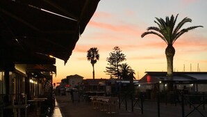 Ein Außenbereich mit Tischen und Stühlen unter einer Überdachung, eine Palme und ein Sonnenuntergang im Hintergrund.