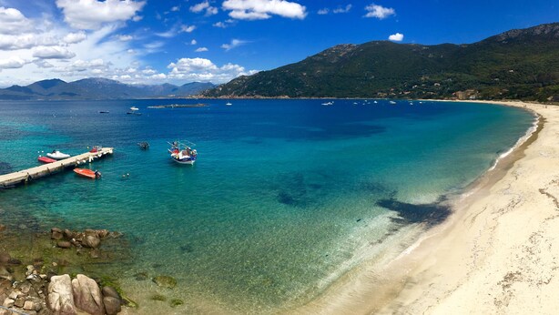 Una playa costera con aguas turquesas cristalinas, barcos amarrados en un muelle y montañas al fondo.