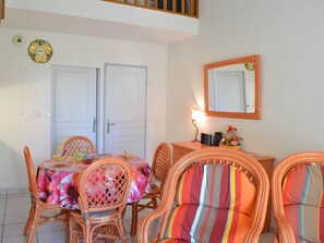 Uma área de refeições com cadeiras de vime e uma mesa coberta por uma toalha de mesa florida. Há duas portas, um espelho e uma pequena mesa com objetos decorativos.