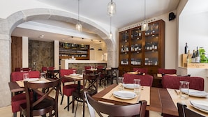 Un restaurant avec des tables en bois, des chaises à l’assise rembourrée de tissu rouge, un présentoir à bouteilles de vin et un mur en pierre.