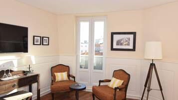 A hotel room with two armchairs, a small table, a desk with a lamp, a floor lamp, a TV, and a window with a view of buildings.