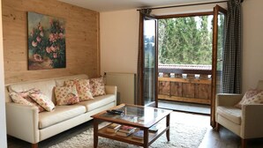 A living room with a sofa, a coffee table with books, a painting on the wall, and a view through a glass door to an outdoor balcony. - Kraeuterhof-Ferienwohnung (Missen-Wilhams)