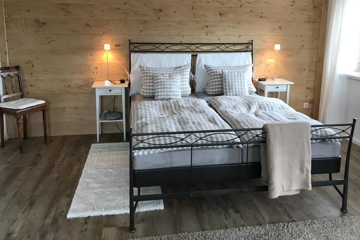 Un dormitorio con pared de madera, estructura de cama metálica, dos mesitas de noche con lámparas, una silla y un ventanal.