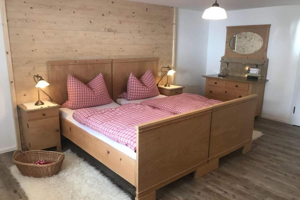 Una cama de madera con una colcha a cuadros, dos mesitas de noche con lámparas, un espejo sobre el tocador y una alfombra mullida en el suelo.