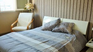 A bed with a patterned bedspread, a white chair, and a vase with a flower.