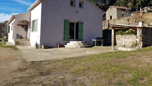 A white building with green shutters, a small porch, and a stone structure nearby.
