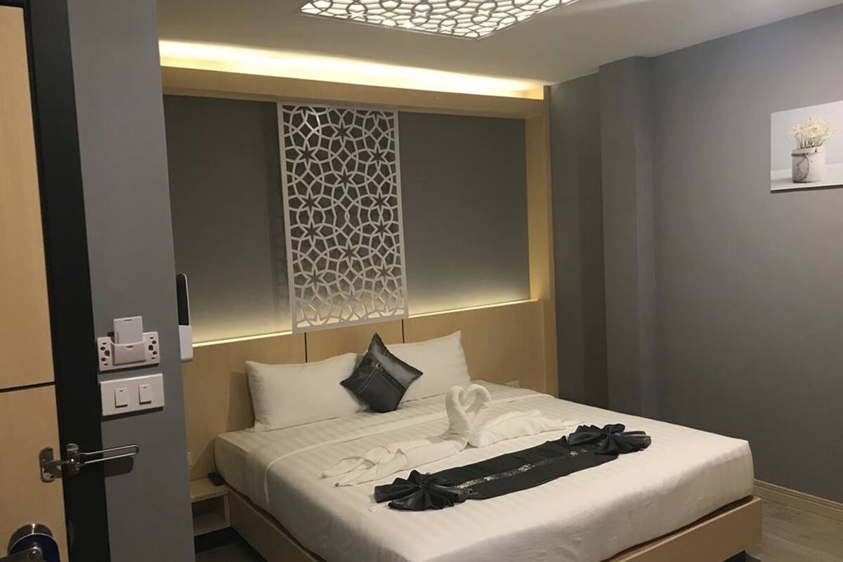 Quarto de hotel moderno, com cama bem arrumada, cabeceira decorativa e luminária fixada na parede.