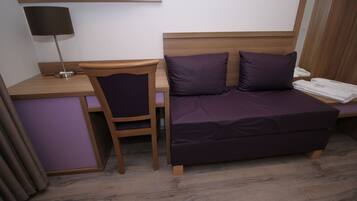 Quarto de hotel compacto com escrivaninha de madeira, uma cadeira, cama com roupa de cama roxa, abajur e televisão fixada na parede.