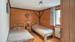 Un dormitorio de cabaña de madera con dos camas individuales, una mesita de noche con una lámpara, una decoración con forma de corazón en la pared y una ventana con cortinas.
