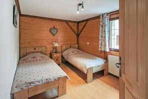 A wooden cabin bedroom with two single beds, a nightstand with a lamp, a heart-shaped decoration on the wall, and a window with curtains. - Homerez - Chalet in Stosswihr (Stosswihr)