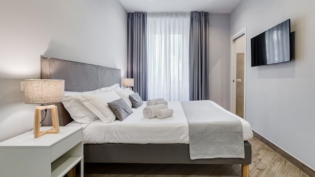 Un dormitorio moderno con cama, mesitas de noche, televisor y ventana con cortinas.