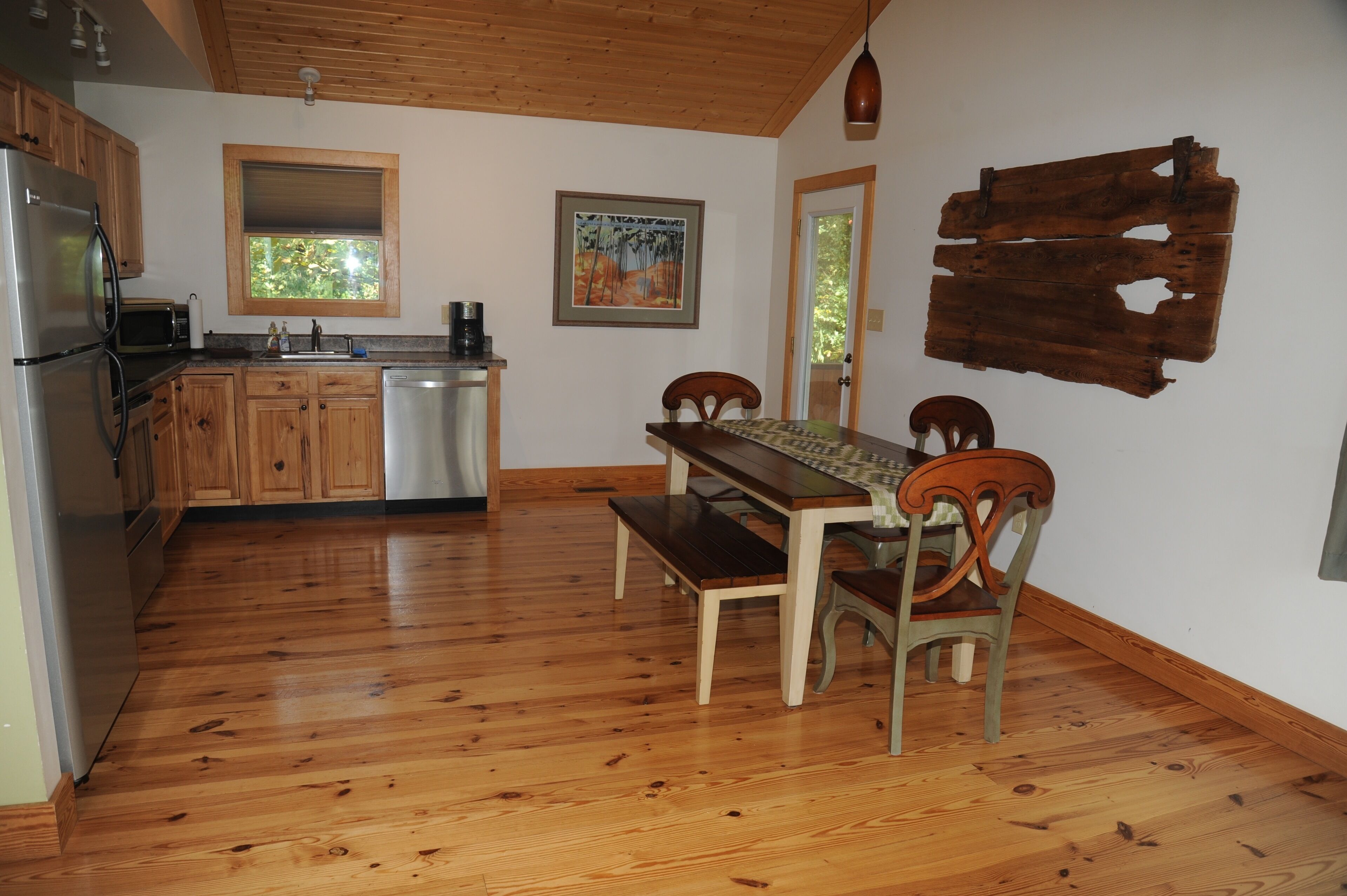 A kitchen with wooden cabinets, a refrigerator, a microwave, and a sink. There is a dining table with chairs and a bench. A painting hangs on the wall, and a wooden board is mounted on the wall.