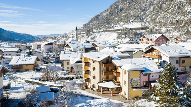 A snow-covered village with traditional Alpine architecture, including multi-story buildings with balconies and a prominent church steeple.