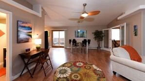 A spacious living room with a ceiling fan, a dining area with a table and chairs, and a sofa with an orange throw.