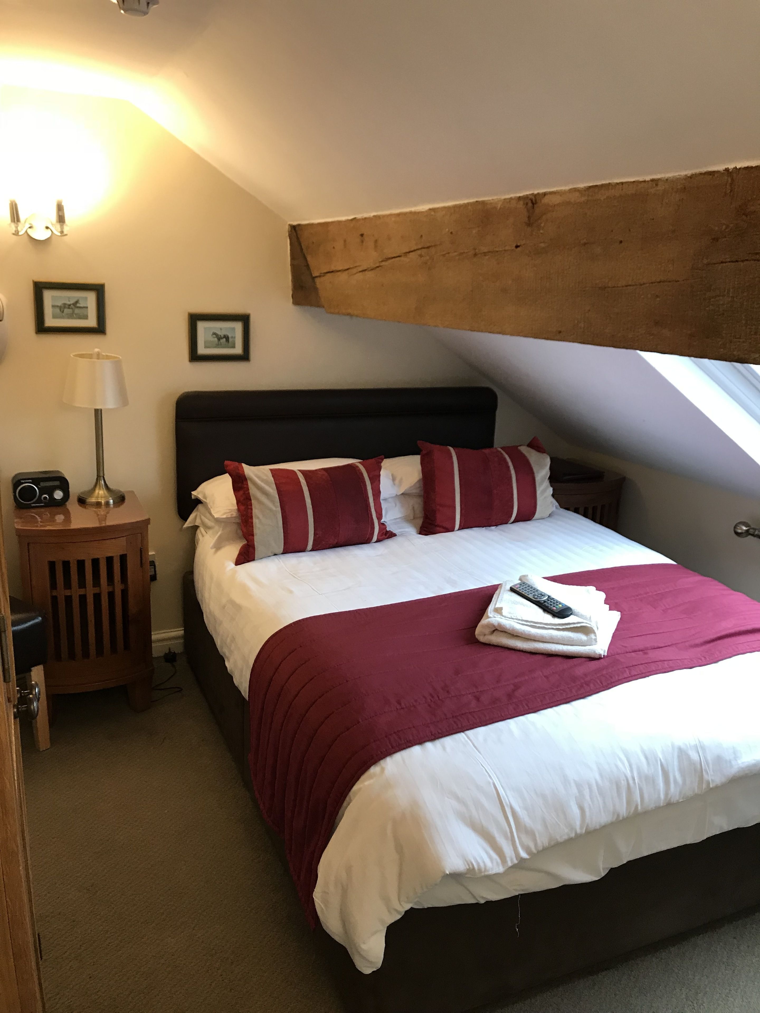 Een net opgemaakt bed met een rode en witte gestreepte kussen, een witte deken en een afstandsbediening erop. Er staat een houten nachtkastje met een lamp en een radio naast het bed.