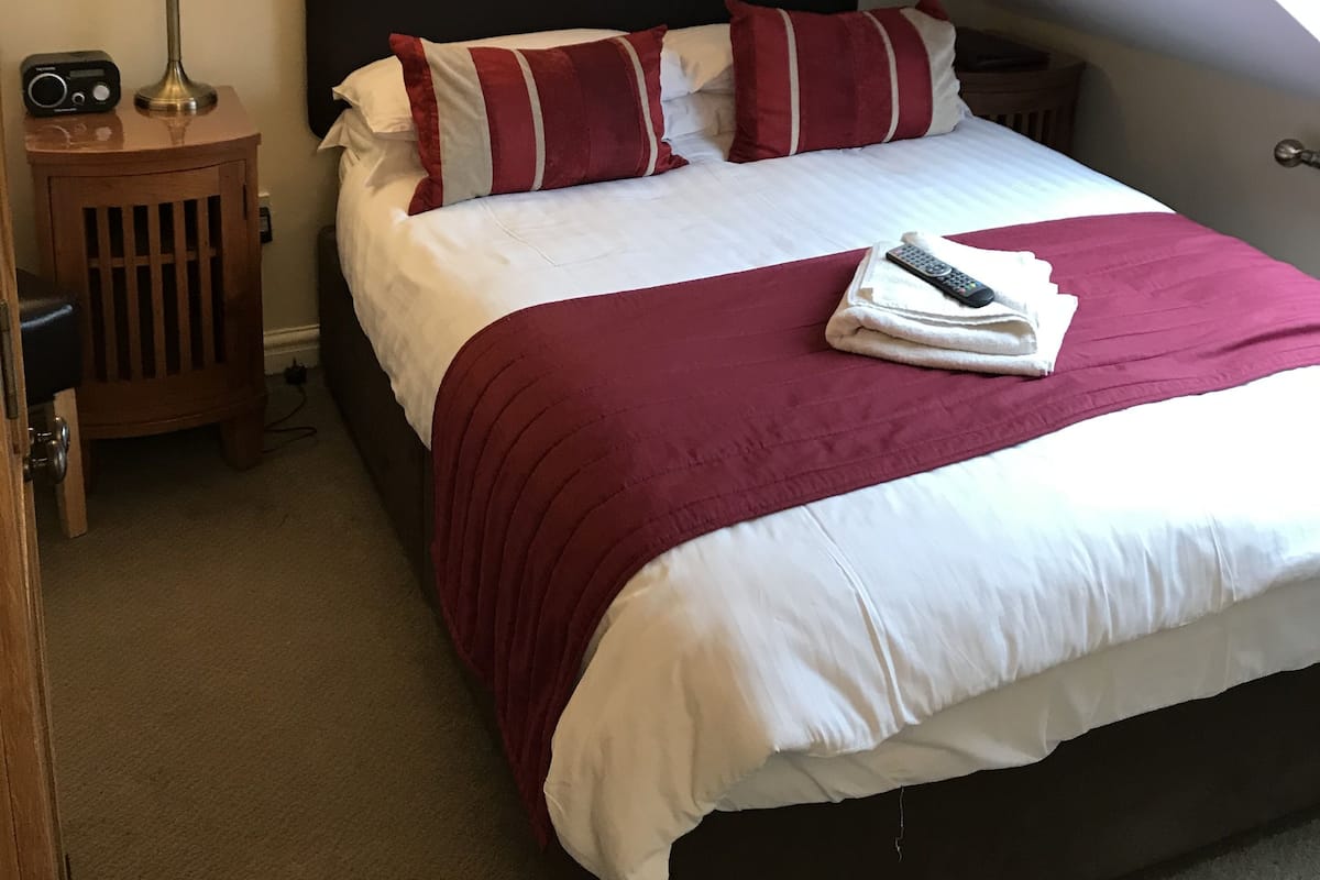 Una cama bien hecha con un cojín a rayas rojas y blancas, una manta blanca y un control remoto encima. Junto a la cama, una mesita de noche de madera con una lámpara y una radio.