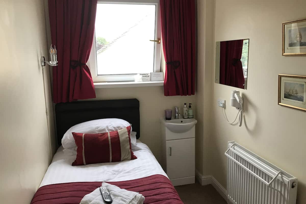 Une petite chambre d’hôtel avec un lit simple, une table de chevet avec une lampe, un radiateur, un petit lavabo et un téléphone.