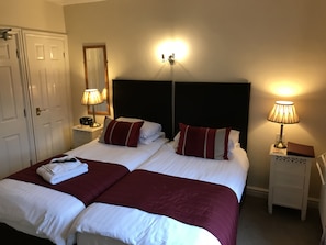 A double bed with two pillows, a bedside table with a lamp, a nightstand with a lamp, and a door.