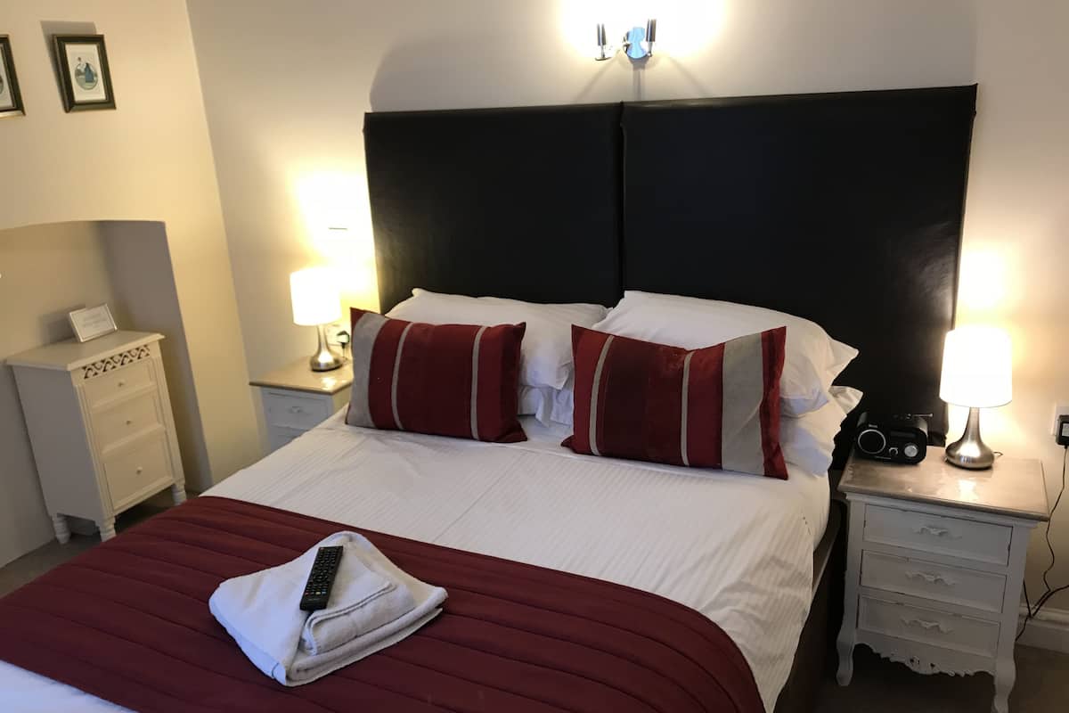 Un lit bien fait, avec un oreiller à rayures rouges et blanches, une télécommande et une serviette pliée posées sur un couvre-lit rouge. On y trouve deux tables de chevet avec des lampes, une petite table d’appoint et un tableau encadré accroché au mur.
