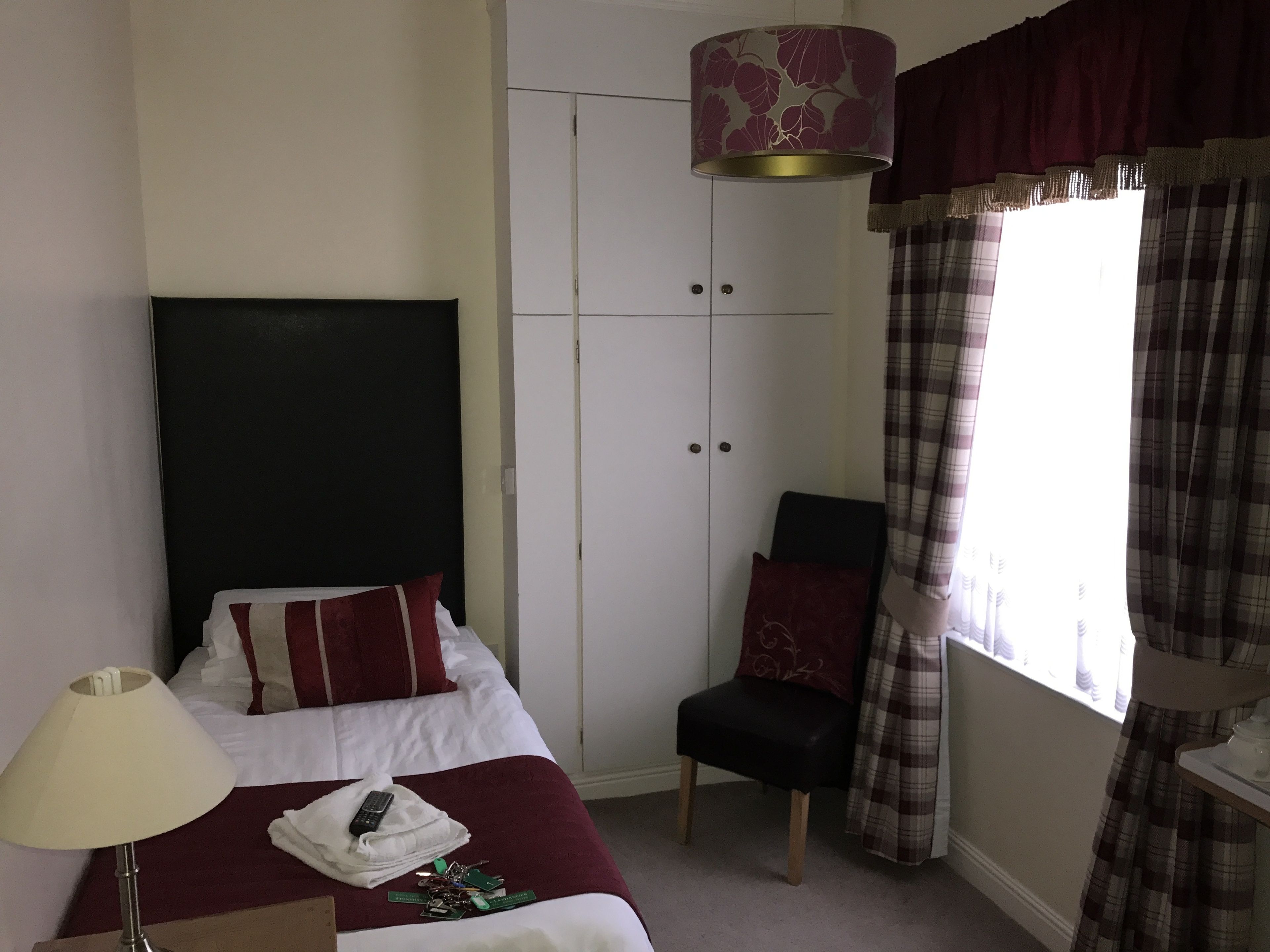 A hotel room with a bed, a chair, a wardrobe, and a window with curtains.
