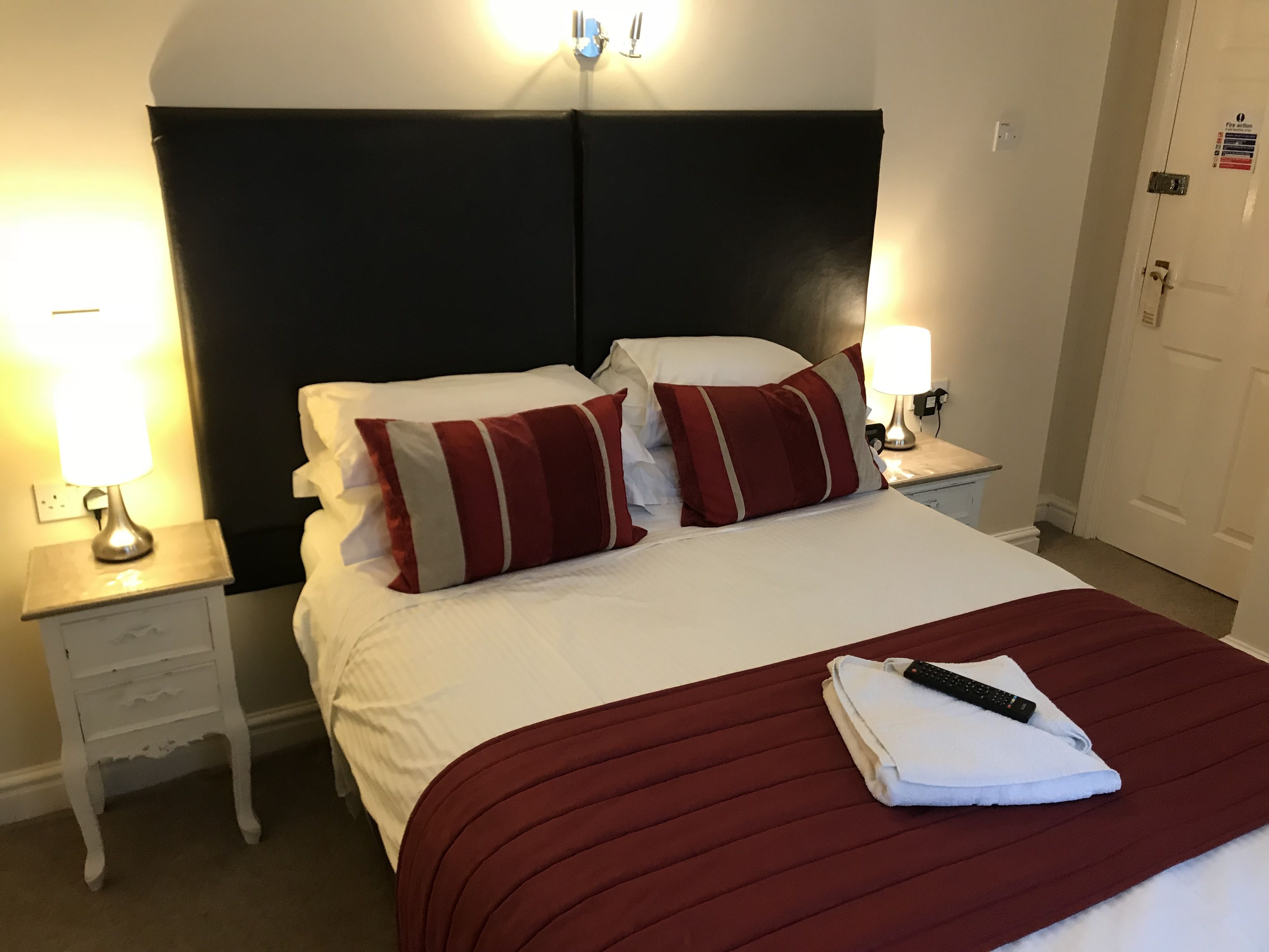Un lit bien fait, avec un oreiller à rayures rouges et blanches, une télécommande et une serviette pliée posées sur un couvre-lit blanc. On y trouve deux tables de chevet avec des lampes, un mur blanc et une porte ornée d’un autocollant indiquant la présence d’un détecteur d’incendie.