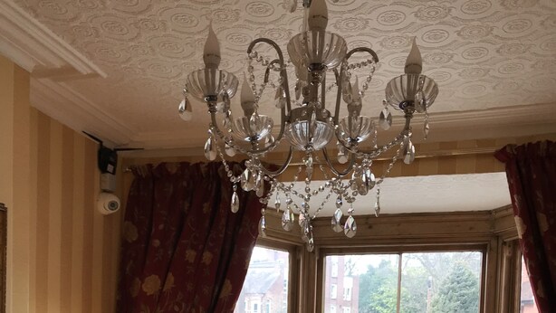 A room with a chandelier, a window with curtains, a lamp, a small table with a bowl, and a sofa.