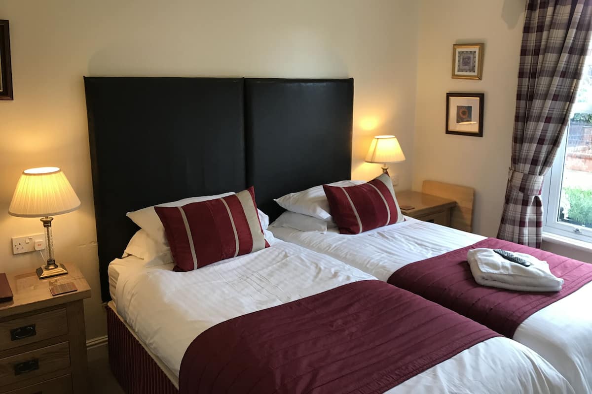 Une chambre d’hôtel avec deux lits, chacun agrémenté de draps blancs et de touches de couleur rouge, d’une tête de lit sombre, de lampes de chevet, d’une table de chevet en bois et d’une fenêtre avec des rideaux.