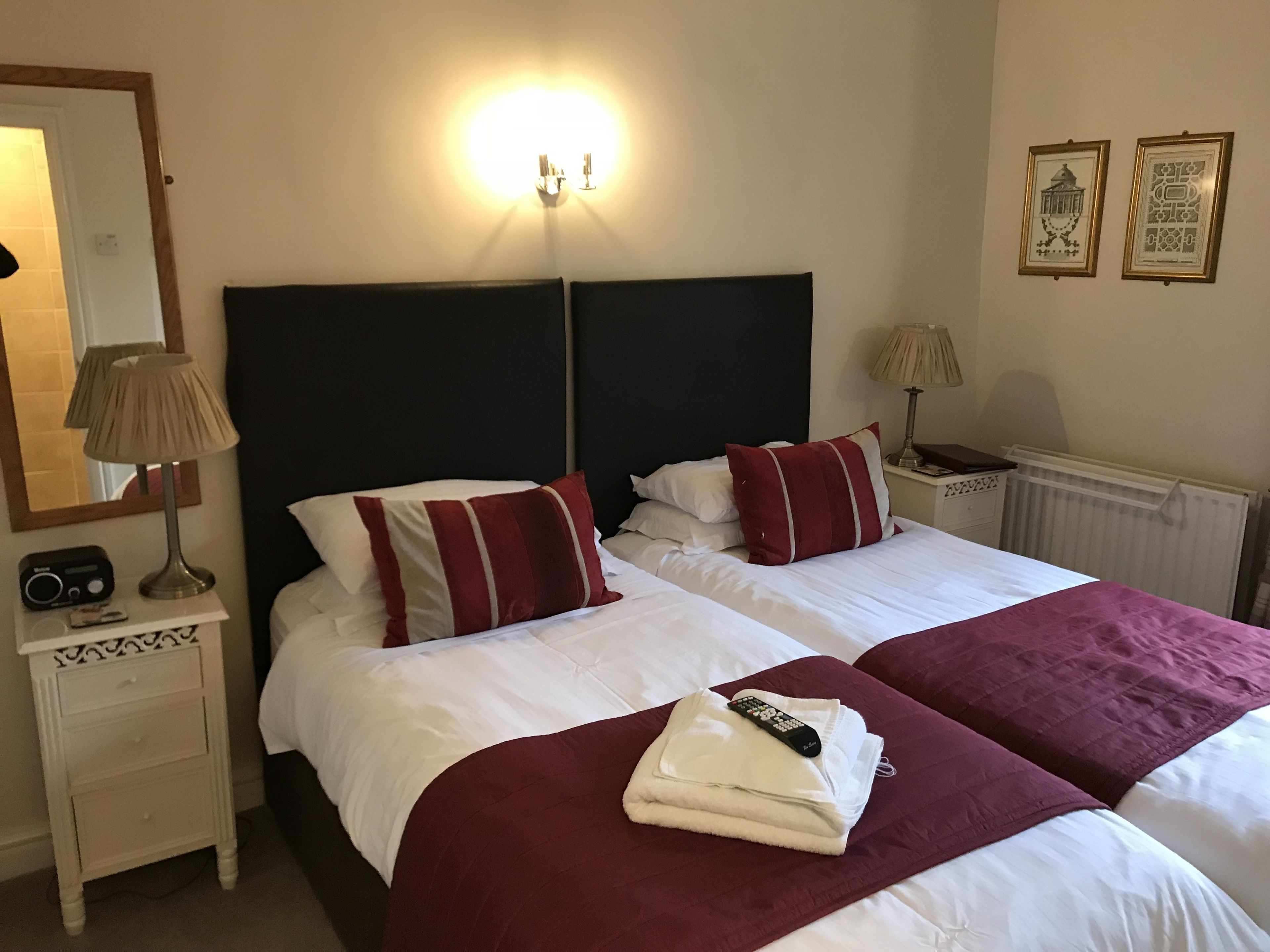 A hotel room with two beds, each with white bedding and red accents, a bedside table with a lamp, a radio, and a framed picture on the wall.