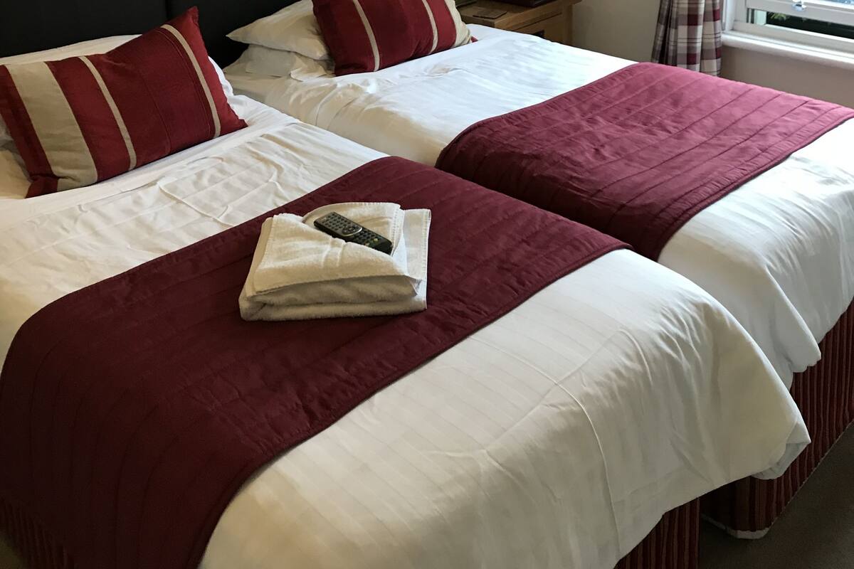 Un lit double avec une literie blanche et rouge, une télécommande sur le lit, une table de chevet avec une lampe et une fenêtre avec des rideaux.